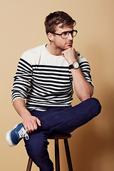 Image showing Thinking, fashion and glasses with a man on a chair in studio on a tan background for vision or contemplation. Future, idea and pose with a confident young person sitting on stool feeling thoughtful