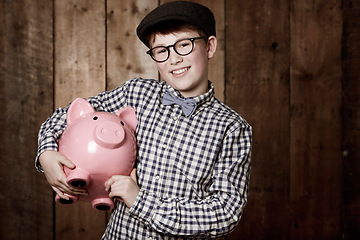 Image showing Young child, piggy bank or smile in portrait by wooden background, money saving or growth in youth in retro fashion. Boy, smile and face for coin banking and safe for bills with glasses by backdrop