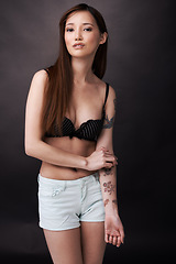 Image showing Asian, woman and serious portrait with tattoos in underwear on dark background of studio. Japanese, model and art with ink on body with lady in jeans, shorts and bra to show unique style or skin