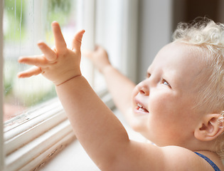 Image showing Baby, rain and happy child by window, curious and smile alone with wet weather. Young kid, glass and dream of blonde toddler thinking of childhood, cute girl playing and watch for learning at house