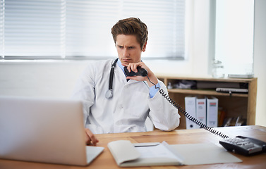 Image showing Laptop, phone call and man doctor in his office for telehealth consultation at hospital. Confused, professional and young male healthcare worker on mobile discussion with landline in medical clinic