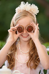 Image showing Child, cupcakes and eyes or play smile or dress up in garden for birthday celebration, dessert or event. Female person, fancy accessories and funny face with sweet food or outdoor, quirky or humor