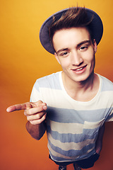 Image showing Portrait, hipster and man with fashion, pointing and confident guy on an orange studio background. Person, hand gesture and model with stylish clothes, hat and casual outfit with aesthetic and trendy