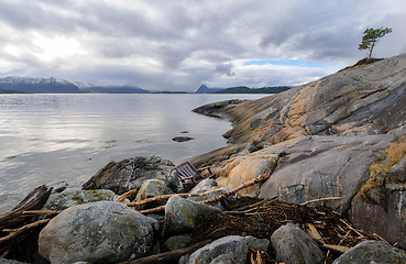 Image showing Rocky shore with driftwood and a a chair that has drifted ashore facing the tranquil sea with mountains beyond.