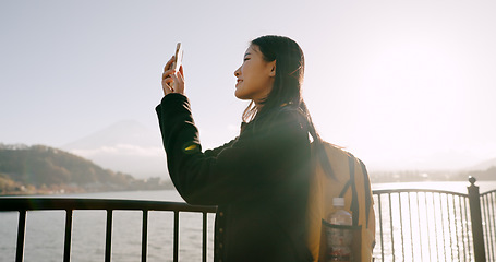 Image showing Japanese woman, lake and phone for photography, memory and post on web blog for travel with backpack. Girl, person and outdoor with smartphone by water, river and photoshoot for social media in Tokyo
