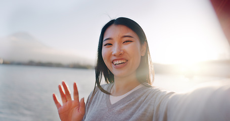 Image showing Japanese woman, selfie and happy outdoor, pointing at lake or river for social media and travel influencer in nature. Live streaming, adventure and wave at camera in Japan with smile in portrait