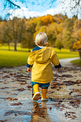 Image showing Sun always shines after the rain. Small bond infant boy wearing yellow rubber boots and yellow waterproof raincoat walking in puddles in city park on sunny rainy day.