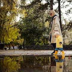 Image showing Small bond infant boy wearing yellow rubber boots and yellow waterproof raincoat walking in puddles on a overcast rainy day holding her mother's hand. Mom with small child in rain outdoors.