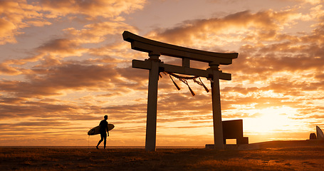 Image showing Torii gate, sunset and man with surfboard, ocean and travel adventure in Japan with orange sky. Shinto architecture, Asian culture and calm beach in Japanese nature with person at spiritual monument.