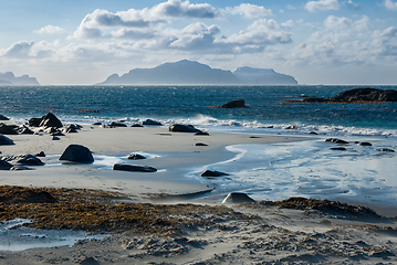 Image showing Calm waters wash over a sandy beach with rocks and seaweed, island silhouette in the distance.