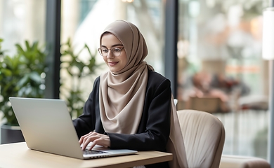 Image showing In a cozy cafe, a woman in a hijab embraces modernity as she diligently works on her laptop, seamlessly blending technology with tradition in a contemporary and culturally diverse workspace