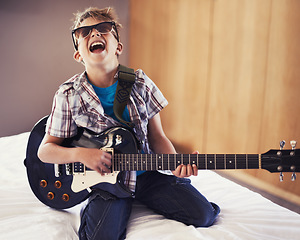 Image showing Happy, playing and child with guitar on bed for music lesson with sunglasses at modern home. Fun, rockstar and young boy kid learning with electric string instrument in bedroom for hobby at house.