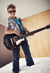 Image showing Smile, playing and kid with guitar on bed for music lesson with sunglasses at modern home. Fun, rockstar and young boy child learning with electric string instrument in bedroom for hobby at house.