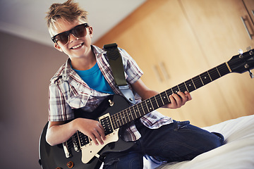 Image showing Smile, playing and child with guitar on bed for music lesson with sunglasses at modern home. Fun, rockstar and young boy kid learning with electric string instrument in bedroom for hobby at house.