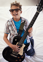 Image showing Happy, playing and boy kid with guitar on bed for music lesson with sunglasses at modern home. Fun, rockstar and young child learning with electric string instrument in bedroom for hobby at house.