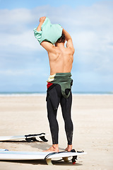 Image showing Surfing, man and getting ready with surfboard on beach with wetsuit, blue sky and dressing with mock up space. Rear view, person and athlete by ocean for training, surfer workout and extreme sports