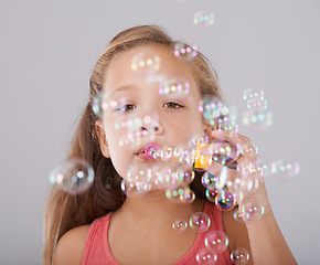 Image showing Portrait, little girl and blowing bubbles in studio by toy, cute and fun games with soap liquid. Child, face and learning to play with bubble wand, childhood development and sweet by gray background