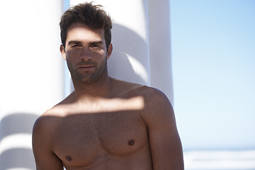 Image showing Handsome, shirtless and portrait of man outdoor by pillars at beach with confidence on vacation. Serious, model and attractive young male person by wall at ocean or sea for holiday or weekend trip.