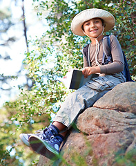 Image showing Boy, kid and hiking by rock and book for reading, backpacking and adventure outdoor. Student on school field trip, relax in nature and forest for discovery or exploration, smile and travel portrait