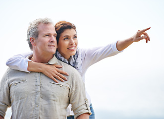 Image showing Happy, mature couple and pointing on holiday with travel to beach and embrace with love on adventure in summer. Vacation, sightseeing and woman hug man in nature on blue sky with mock up space