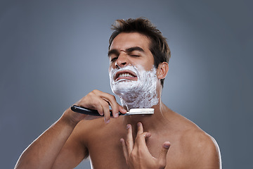 Image showing Man, shaving and razor with stress in studio for grooming, skincare and wellness with pain by grey background. Person, model and annoyed with beard, facial hair removal and cream for cosmetic change