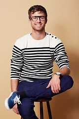 Image showing Portrait, fashion and glasses with a man on a chair in studio on tan background for trendy style. Relax, casual and eyewear with a confident young geek or nerd sitting on a stool in a clothes outfit