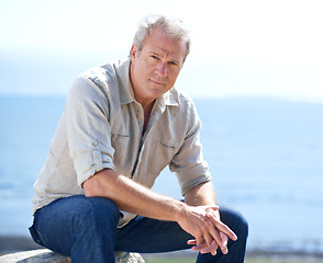 Image showing Mature, man and outdoor portrait at the sea on holiday, vacation or travel in nature to relax. Serious, person and calm ocean, water or waves in morning and sitting on a rock with blue sky in summer