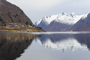 Image showing A tranquil fjord with a village on the shore, reflecting snow-covered mountains in calm water.