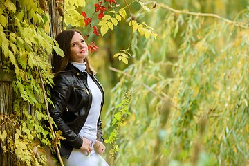 Image showing A young beautiful girl pressed her back against a wooden wall and looks at the autumn forest