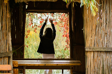 Image showing A young beautiful girl stands in the doorway of an old hut and raised her hands up