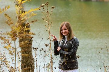 Image showing A girl stands by the lake on a warm autumn day, and points to a plant and smiles happily