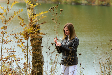 Image showing A girl stands by the lake on a warm autumn day and examines the leaves of plants.