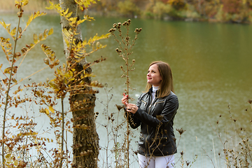Image showing A girl stands by the lake on a warm autumn day and holds a tall dry bush