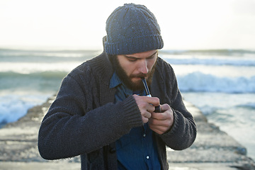 Image showing Bearded, man and smoking a pipe by ocean, lighter and tobacco habit on winter morning for sunrise. English guy, nicotine and vintage smoker for calm, satisfaction and vacation on beach in cape town