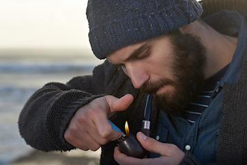 Image showing Bearded, man and smoking a pipe on beach, lighter and tobacco habit on winter morning for sunrise. English guy, nicotine and vintage smoker for calm, satisfaction and vacation by ocean in cape town