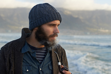 Image showing Beard, man and smoking a pipe by ocean, thinking and sailor habit on winter morning to relax. English person, nicotine and vintage smoker with breathe smoke, calm and vacation in cape town by beach