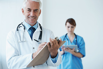 Image showing Portrait, mature doctor writing on his clipboard and with a nurse browsing on a digital tablet in the background. Senior doctor and nurse standing in corridor looking at files discussing reports