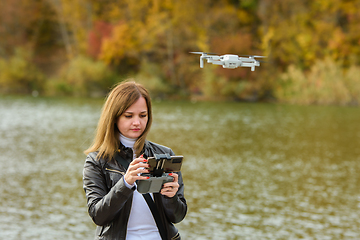 Image showing A young beautiful girl launches a radio-controlled quadcopter on the shore of an autumn lake, the girl adjusts the camera