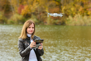 Image showing A young beautiful girl launches a radio-controlled quadcopter on the shore of an autumn lake, the girl looks at the quadcopter