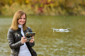 Image showing A young beautiful girl launches a radio-controlled quadcopter on the shore of an autumn lake, the girl looks at the drone smiling