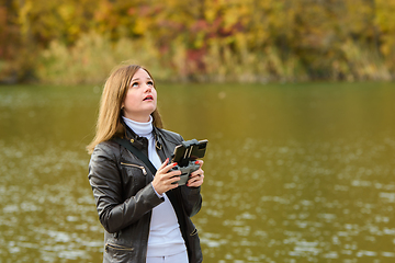 Image showing A young beautiful girl launches a radio-controlled quadcopter on the shore of an autumn lake, the girl looks above herself in search of a drone
