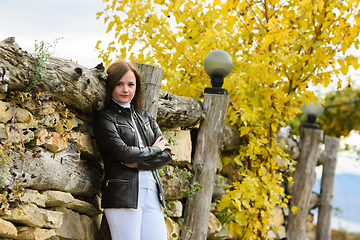 Image showing Young beautiful girl in casual clothes against the background of an old fence and autumn yellow foliage