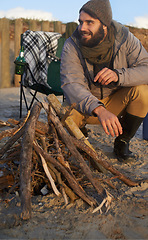 Image showing Campfire, wood and man by beach on vacation, adventure or holiday camping for travel. Nature, outdoor and young male person sitting on chair in sand with flame for heat on weekend trip in winter.