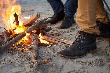 Image showing Closeup, campfire and friends at the beach on holiday, vacation and travel to warm up in winter outdoor. Flame, wood and people together by sand at seashore with shoes, adventure and legs in nature