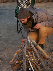 Image showing Campfire, nature and man by beach for travel on vacation, adventure or holiday camping. Wood, outdoor and young male person sitting on chair in sand with flame for heat on weekend trip in winter.