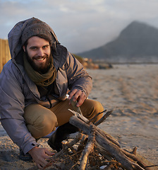 Image showing Portrait, smile and man at beach with campfire on holiday, vacation or travel toe prepare for winter outdoor alone. Happy person, wood and adventure at seashore, leisure and recreation in Canada