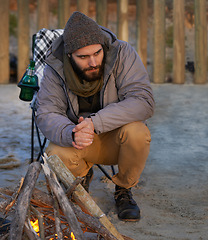 Image showing Campfire, wood and man by beach for travel on vacation, adventure or holiday camping. Nature, outdoor and young male person sitting on chair in sand with flame for heat on weekend trip in winter.