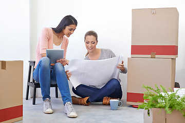 Image showing Women, blueprint and talking on floor at startup with tablet, idea or entrepreneurship with planning. People, partnership and digital touchscreen by boxes for new business with documents at company