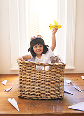 Image showing Child, smile and airplane toy in home, playful and freedom of youth with paper for fun and games. Little kid, happiness and hand in air with body in basket with hat on head as helmet with imagination
