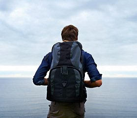 Image showing Man, backpack and hiking by ocean for travel, sightseeing or outdoor journey in nature. Rear view of male person, hiker or tourist with bag on back for trekking, fitness or adventure with cloudy sky